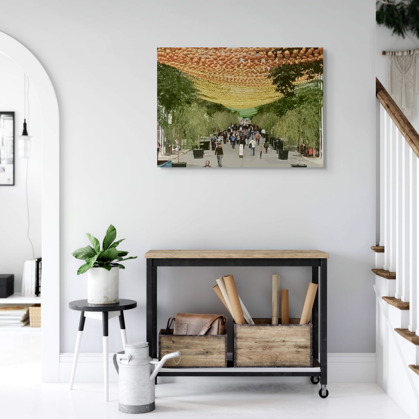 The Village Canvas Wall Art in home foyer with white wall, staircase, wooden entry table, two wooden boxes, black side table and pot planet