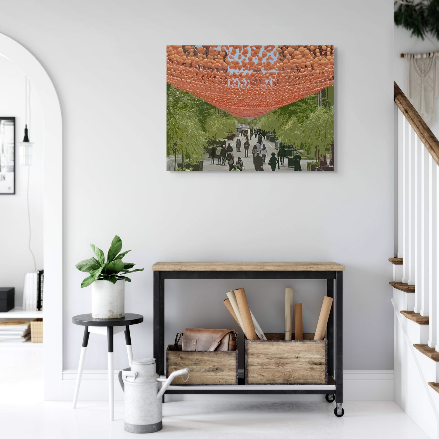 The Village Canvas Wall Art in home foyer with white wall, staircase, wooden entry table, two wooden boxes, black side table and pot plant